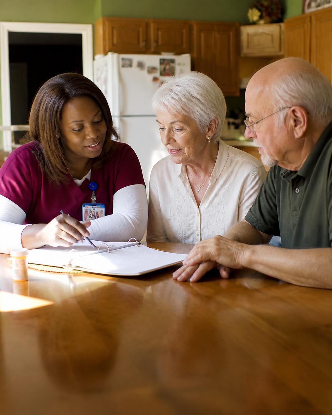 A nursing assistant in scrubs reviewing a care plan with an elderly couple at their kitchen table